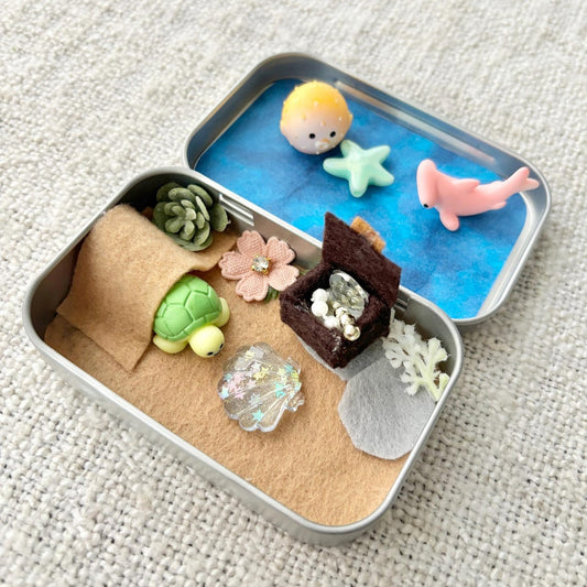 Miniature diorama in a small metal container with various small figurines on a textured surface