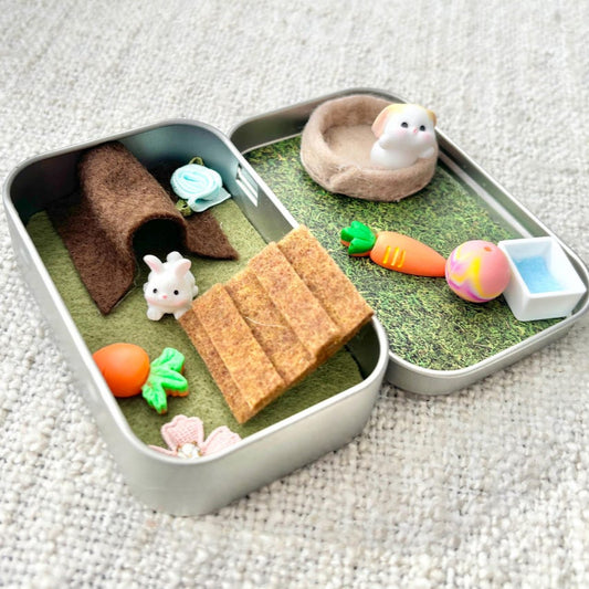 Small play setup with tunnels, toys, and grass-like material inside a tin on a textured surface.