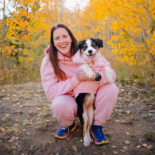 Woman in pink outfit holding a dog with autumn trees in the background